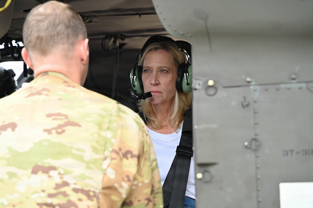 A civilian woman wearing green aviation headset and a shoulder harness is seated inside what appears to be a military helicopter, engaged in conversation with a uniformed soldier in MultiCam/OCP camouflage. The setting suggests a briefing, orientation, or familiarization visit to a military aircraft. The aircraft exterior shows olive drab metal paneling consistent with a military rotary-wing platform.