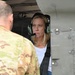 A civilian woman wearing green aviation headset and a shoulder harness is seated inside what appears to be a military helicopter, engaged in conversation with a uniformed soldier in MultiCam/OCP camouflage. The setting suggests a briefing, orientation, or familiarization visit to a military aircraft. The aircraft exterior shows olive drab metal paneling consistent with a military rotary-wing platform.