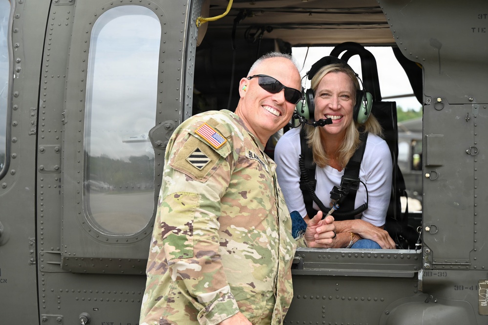 A U.S. Army soldier in OCP (Operational Camouflage Pattern) uniform poses with a civilian woman who is seated inside a military helicopter wearing aviation headgear. The soldier displays a 3rd Infantry Division patch and an American flag on their sleeve. Both individuals are smiling, suggesting a positive, informal interaction likely during a demonstration or familiarization event.