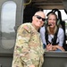 A U.S. Army soldier in OCP (Operational Camouflage Pattern) uniform poses with a civilian woman who is seated inside a military helicopter wearing aviation headgear. The soldier displays a 3rd Infantry Division patch and an American flag on their sleeve. Both individuals are smiling, suggesting a positive, informal interaction likely during a demonstration or familiarization event.