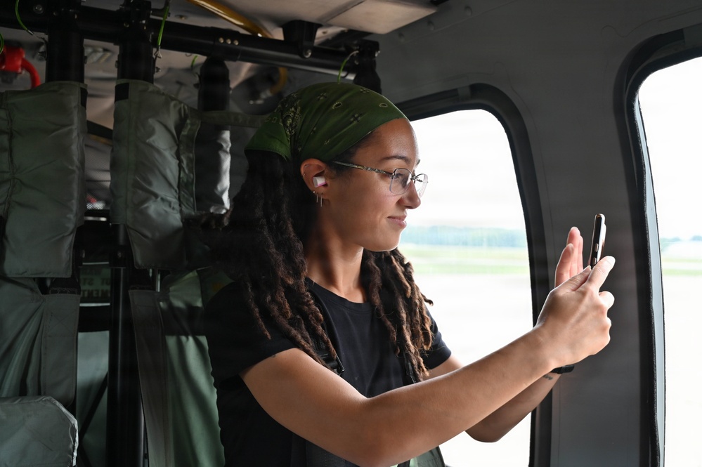 A young woman with dreadlocks and a green bandana is seated inside what appears to be a military or utility helicopter, taking a photo or video with her smartphone through the window. She is wearing glasses, a black t-shirt, and has wireless earbuds in her ears. The interior of the aircraft shows olive-green equipment bags and structural components typical of a military rotary-wing aircraft.