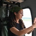 A young woman with dreadlocks and a green bandana is seated inside what appears to be a military or utility helicopter, taking a photo or video with her smartphone through the window. She is wearing glasses, a black t-shirt, and has wireless earbuds in her ears. The interior of the aircraft shows olive-green equipment bags and structural components typical of a military rotary-wing aircraft.
