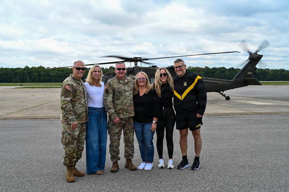Six individuals pose for a group photo on a military airfield tarmac in front of a UH-60 Black Hawk helicopter with its rotors spinning. The group includes two soldiers in Army Combat Uniforms (ACU/OCP pattern), one person in Army PT (physical training) attire, and three civilians. The setting appears to be a formal or commemorative occasion at a military installation.