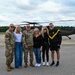 Six individuals pose for a group photo on a military airfield tarmac in front of a UH-60 Black Hawk helicopter with its rotors spinning. The group includes two soldiers in Army Combat Uniforms (ACU/OCP pattern), one person in Army PT (physical training) attire, and three civilians. The setting appears to be a formal or commemorative occasion at a military installation.