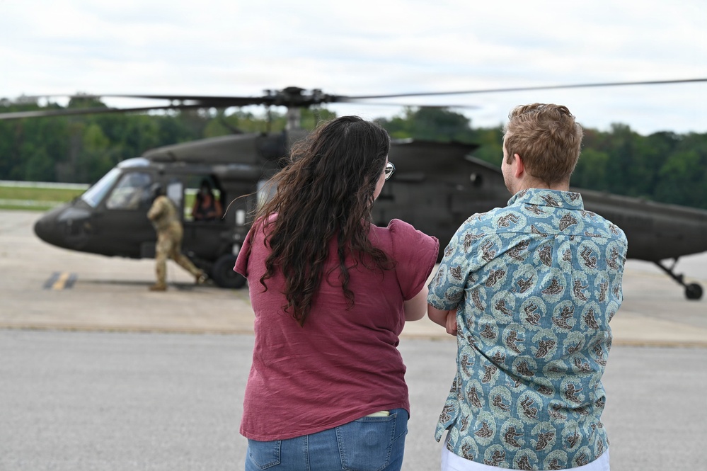 Two civilians stand on a tarmac observing a military helicopter, likely a UH-60 Black Hawk, at close range. A uniformed military personnel member is visible near the aircraft's door. The scene appears to be a public event, demonstration, or family day at a military installation.