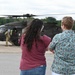 Two civilians stand on a tarmac observing a military helicopter, likely a UH-60 Black Hawk, at close range. A uniformed military personnel member is visible near the aircraft's door. The scene appears to be a public event, demonstration, or family day at a military installation.