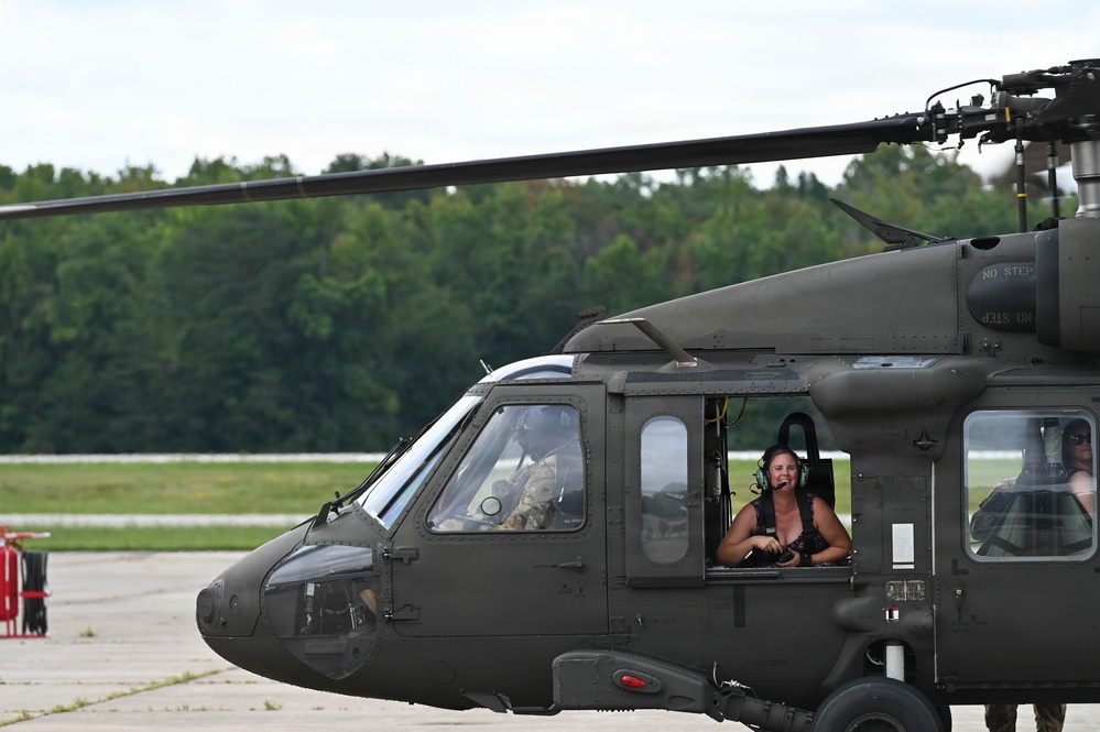 A military UH-60 Black Hawk helicopter is shown on a tarmac or airfield, with multiple occupants visible through the cockpit and side windows. A smiling woman wearing a green flight helmet and headset is prominently visible through the open side door window. The helicopter is painted in standard olive drab military finish and appears to be on the ground at a military installation or airshow.