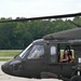 A military UH-60 Black Hawk helicopter is shown on a tarmac or airfield, with multiple occupants visible through the cockpit and side windows. A smiling woman wearing a green flight helmet and headset is prominently visible through the open side door window. The helicopter is painted in standard olive drab military finish and appears to be on the ground at a military installation or airshow.