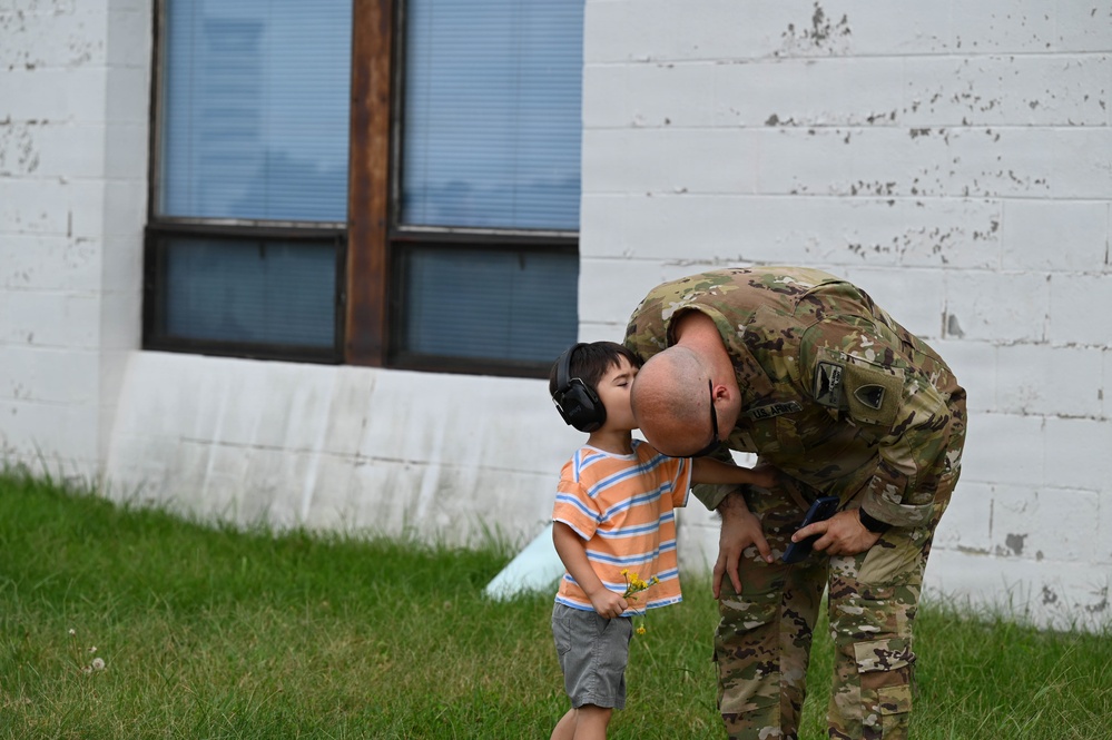 A U.S. Army soldier in OCP (Operational Camouflage Pattern) uniform bends down to share a kiss or forehead touch with a young child wearing ear protection headphones. The child is dressed in a striped shirt and shorts and appears to be holding a small flower. The scene takes place on a grass lawn in front of a white-painted concrete block building.