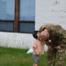A U.S. Army soldier in OCP (Operational Camouflage Pattern) uniform bends down to share a kiss or forehead touch with a young child wearing ear protection headphones. The child is dressed in a striped shirt and shorts and appears to be holding a small flower. The scene takes place on a grass lawn in front of a white-painted concrete block building.