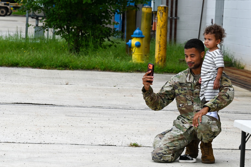 A soldier in U.S. Army OCP (Operational Camouflage Pattern) uniform, with the name tape 'MITCHELL' visible, kneels on a concrete surface while holding a young toddler on his knee and taking a photo or video with a smartphone. The setting appears to be a military installation or industrial area, with a yellow fire hydrant, yellow bollards, and a blue building visible in the background. The moment appears to capture a family reunion or casual interaction during a military event.