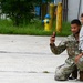 A soldier in U.S. Army OCP (Operational Camouflage Pattern) uniform, with the name tape 'MITCHELL' visible, kneels on a concrete surface while holding a young toddler on his knee and taking a photo or video with a smartphone. The setting appears to be a military installation or industrial area, with a yellow fire hydrant, yellow bollards, and a blue building visible in the background. The moment appears to capture a family reunion or casual interaction during a military event.