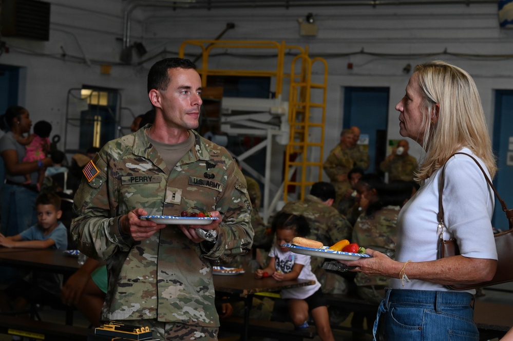 A U.S. Army soldier named Perry is engaged in conversation with a civilian woman during what appears to be a unit family gathering or cookout event. Both individuals are holding food plates, suggesting a casual social meal setting inside a military facility or hangar. Other soldiers and children are visible in the background, reinforcing the family-oriented nature of the event.