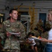 A U.S. Army soldier named Perry is engaged in conversation with a civilian woman during what appears to be a unit family gathering or cookout event. Both individuals are holding food plates, suggesting a casual social meal setting inside a military facility or hangar. Other soldiers and children are visible in the background, reinforcing the family-oriented nature of the event.