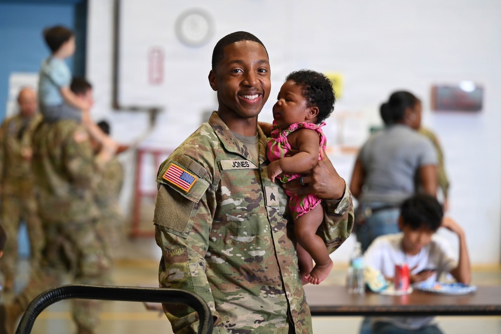 A U.S. Army Sergeant named Jones, wearing OCP (Operational Camouflage Pattern) uniform, smiles broadly while holding a young infant dressed in a pink outfit. The setting appears to be a military facility or gymnasium during what looks like a family reunion or unit gathering event. Multiple soldiers and civilians are visible in the background engaged in similar social interactions.
