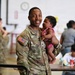 A U.S. Army Sergeant named Jones, wearing OCP (Operational Camouflage Pattern) uniform, smiles broadly while holding a young infant dressed in a pink outfit. The setting appears to be a military facility or gymnasium during what looks like a family reunion or unit gathering event. Multiple soldiers and civilians are visible in the background engaged in similar social interactions.