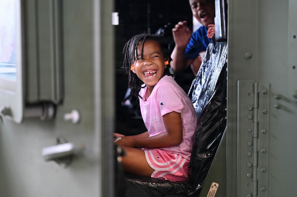 A young girl wearing a pink Champion t-shirt and pink striped shorts is laughing joyfully while seated inside what appears to be a military vehicle, visible through an open green metal door. A second child in a blue shirt is partially visible behind her. The scene captures a candid, high-energy moment of children exploring or riding in the vehicle.