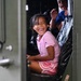 A young girl wearing a pink Champion t-shirt and pink striped shorts is laughing joyfully while seated inside what appears to be a military vehicle, visible through an open green metal door. A second child in a blue shirt is partially visible behind her. The scene captures a candid, high-energy moment of children exploring or riding in the vehicle.