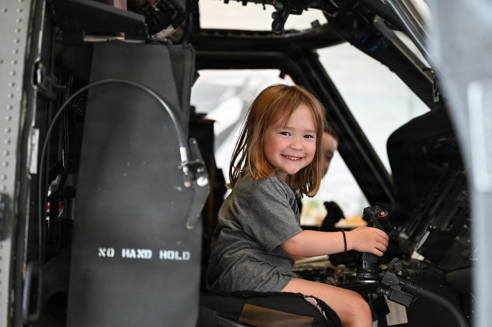 A young girl with reddish-brown hair is seated in the cockpit of a military helicopter, smiling broadly at the camera. She is holding the control stick (cyclic) and appears to be enjoying a public access or airshow event. The cockpit interior is dark-colored with various instruments and controls visible.