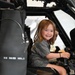 A young girl with reddish-brown hair is seated in the cockpit of a military helicopter, smiling broadly at the camera. She is holding the control stick (cyclic) and appears to be enjoying a public access or airshow event. The cockpit interior is dark-colored with various instruments and controls visible.