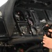 A person's hands are holding a flight control stick (joystick) in what appears to be a military aircraft cockpit. The cockpit features multiple instrument panels, displays, and avionics equipment. A brand label reading 'AVALEX' is visible on one of the display units in the background.