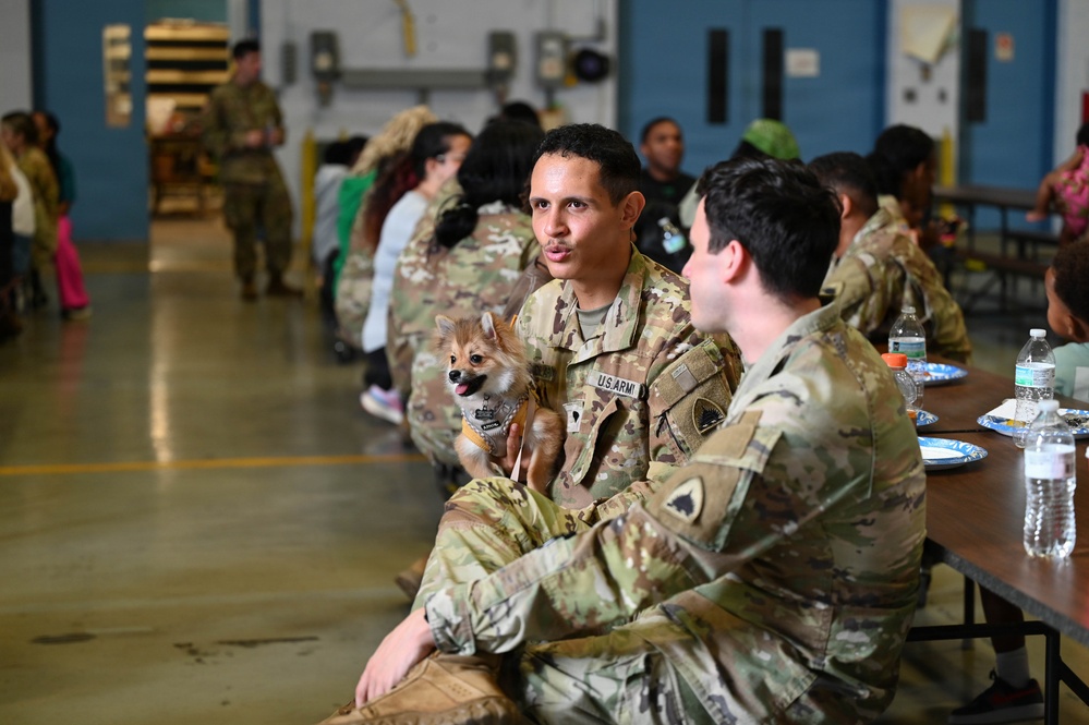 U.S. Army soldiers are gathered in what appears to be a military hangar or large indoor facility, likely during a unit gathering or family day event. A soldier in the foreground is holding a small Pomeranian-type dog while conversing with another soldier. The scene includes civilians, including children, in the background suggesting a family-oriented military event.