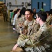 U.S. Army soldiers are gathered in what appears to be a military hangar or large indoor facility, likely during a unit gathering or family day event. A soldier in the foreground is holding a small Pomeranian-type dog while conversing with another soldier. The scene includes civilians, including children, in the background suggesting a family-oriented military event.