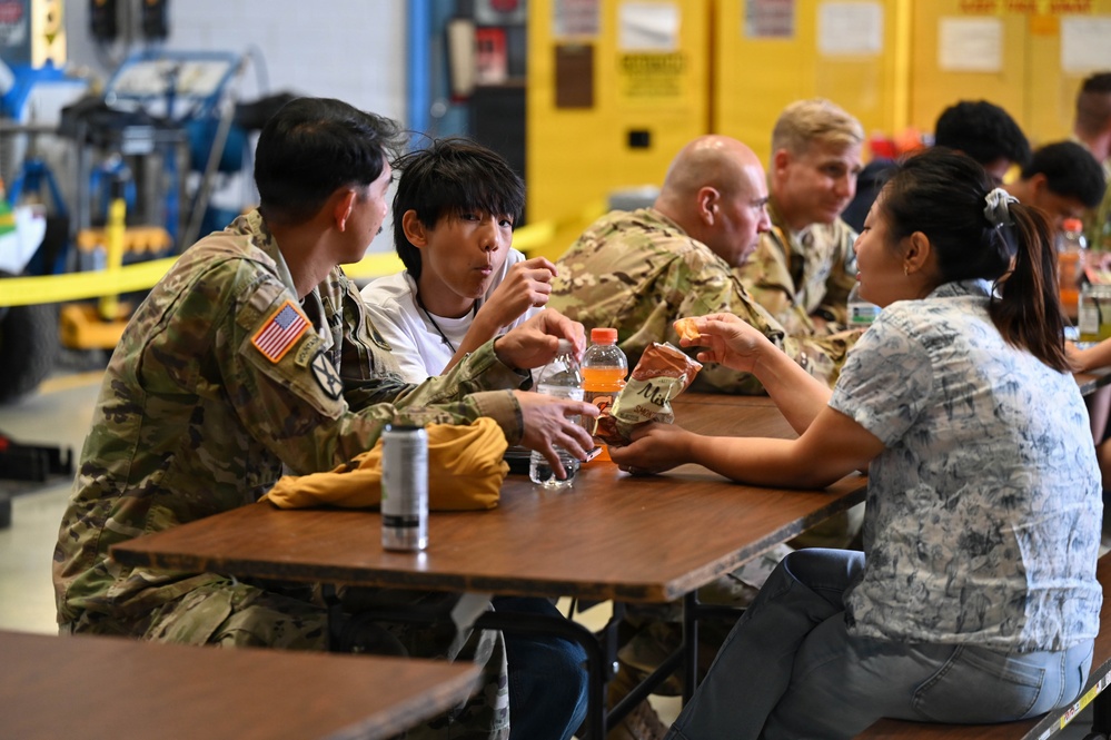 A group of U.S. Army soldiers in MultiCam/OCP camouflage uniforms are seated at tables in what appears to be a military hangar or maintenance facility, sharing a meal with civilians. The scene suggests an informal gathering or community engagement event, with both military personnel and civilians eating together. The background shows yellow industrial equipment and safety markings typical of a military maintenance bay.