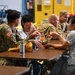 A group of U.S. Army soldiers in MultiCam/OCP camouflage uniforms are seated at tables in what appears to be a military hangar or maintenance facility, sharing a meal with civilians. The scene suggests an informal gathering or community engagement event, with both military personnel and civilians eating together. The background shows yellow industrial equipment and safety markings typical of a military maintenance bay.