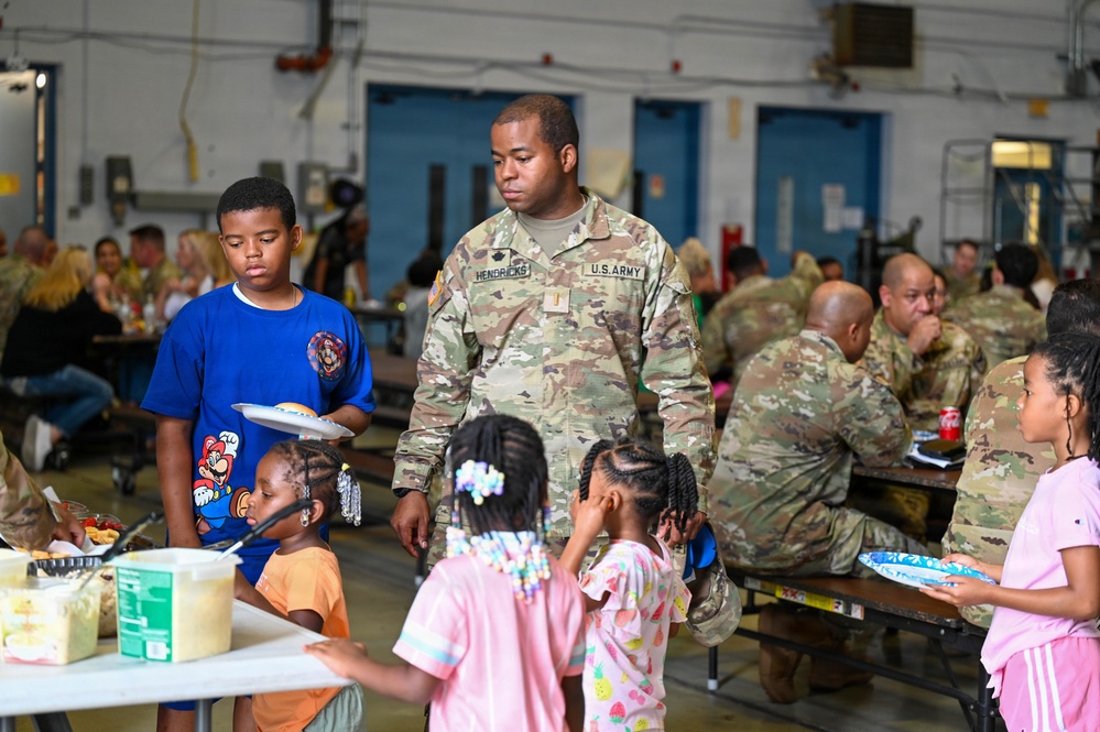 A U.S. Army soldier named Hendricks, wearing OCP (Operational Camouflage Pattern) uniform, is present at what appears to be a military dining facility or mess hall event with several children. The setting suggests a family day or community event at a military installation, with multiple soldiers and civilians gathered at long tables eating. Children of various ages are visible holding plates and moving around the food service area.