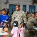 A U.S. Army soldier named Hendricks, wearing OCP (Operational Camouflage Pattern) uniform, is present at what appears to be a military dining facility or mess hall event with several children. The setting suggests a family day or community event at a military installation, with multiple soldiers and civilians gathered at long tables eating. Children of various ages are visible holding plates and moving around the food service area.