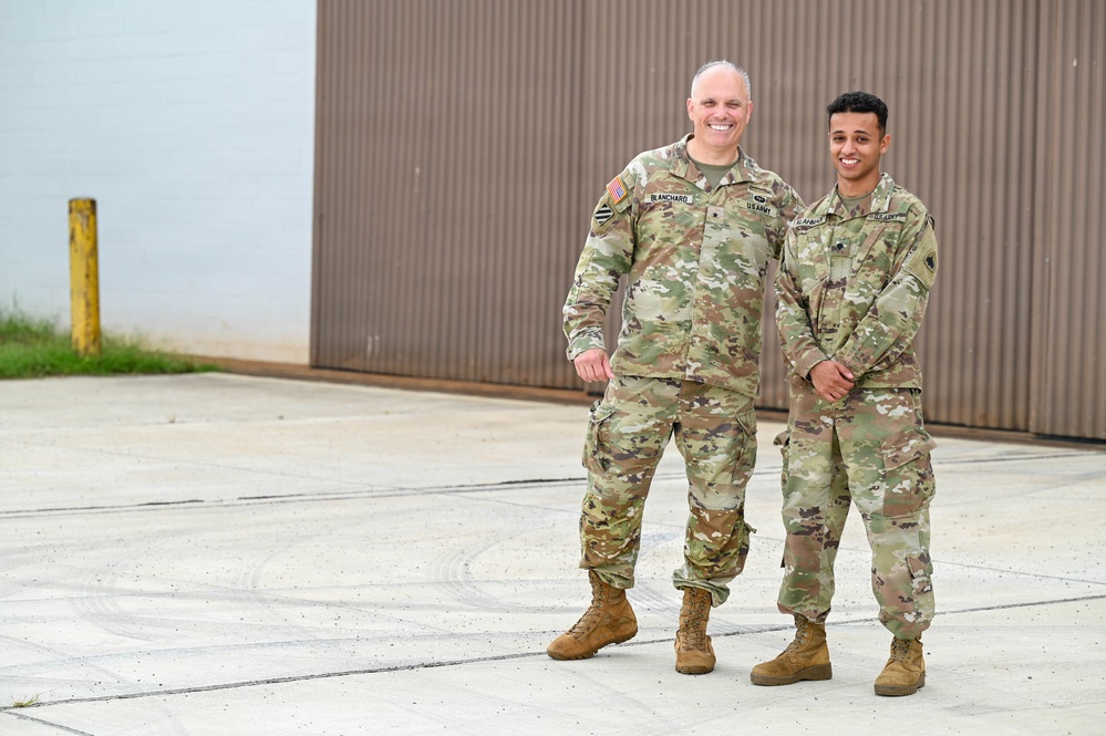 Two U.S. Army soldiers in Operational Camouflage Pattern (OCP) uniforms are posing together outdoors in front of a corrugated metal building. Both are smiling at the camera in what appears to be a formal portrait-style photograph taken on a paved surface. The name tape 'BLANCHARD' is visible on the taller soldier on the left.