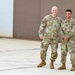 Two U.S. Army soldiers in Operational Camouflage Pattern (OCP) uniforms are posing together outdoors in front of a corrugated metal building. Both are smiling at the camera in what appears to be a formal portrait-style photograph taken on a paved surface. The name tape 'BLANCHARD' is visible on the taller soldier on the left.
