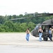 A military UH-60 Black Hawk helicopter is parked on a tarmac or airfield apron. Three civilian women and one uniformed military personnel are visible near the aircraft, suggesting a tour, media visit, or familiarization event. The setting appears to be a military airfield surrounded by dense green treeline.