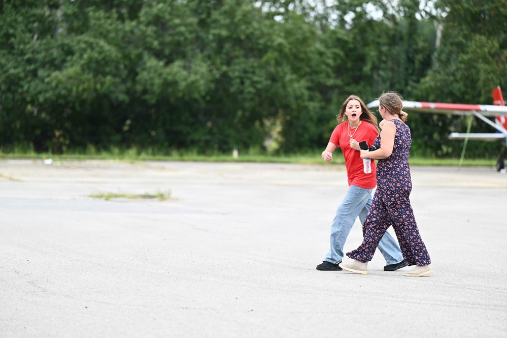 Two young women are walking across a large paved open area, possibly a parking lot or tarmac. The woman in a red t-shirt and jeans appears to be speaking or reacting expressively with her mouth open, while the other woman in a floral jumpsuit walks beside her. A tree line and what appears to be a barrier or gate structure are visible in the background.