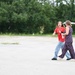 Two young women are walking across a large paved open area, possibly a parking lot or tarmac. The woman in a red t-shirt and jeans appears to be speaking or reacting expressively with her mouth open, while the other woman in a floral jumpsuit walks beside her. A tree line and what appears to be a barrier or gate structure are visible in the background.