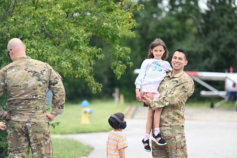 A smiling soldier in U.S. Army OCP (Operational Camouflage Pattern) uniform holds a young girl while a young boy wearing noise-canceling headphones stands nearby. A second soldier with his back to the camera is visible on the left side. The setting appears to be a military airfield or tarmac, with a small aircraft visible in the background.