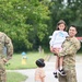 A smiling soldier in U.S. Army OCP (Operational Camouflage Pattern) uniform holds a young girl while a young boy wearing noise-canceling headphones stands nearby. A second soldier with his back to the camera is visible on the left side. The setting appears to be a military airfield or tarmac, with a small aircraft visible in the background.