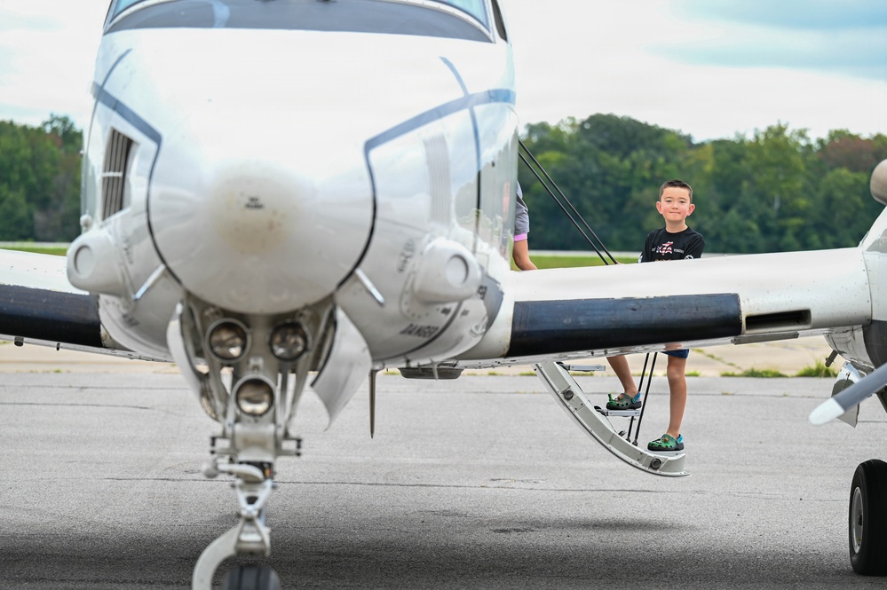 A young boy stands on the wing step of a small white propeller aircraft on a tarmac, smiling toward the camera. The image is taken from the front of the aircraft, with the propeller hub and nose gear prominently in the foreground. The setting appears to be a small general aviation airport with trees visible in the background.