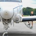 A young boy stands on the wing step of a small white propeller aircraft on a tarmac, smiling toward the camera. The image is taken from the front of the aircraft, with the propeller hub and nose gear prominently in the foreground. The setting appears to be a small general aviation airport with trees visible in the background.