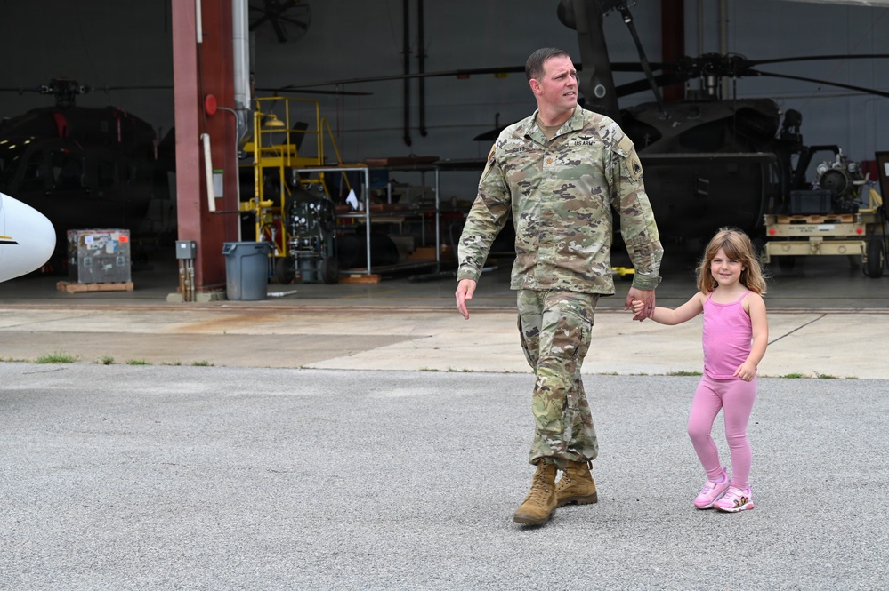 A U.S. Army soldier in OCP (Operational Camouflage Pattern) uniform walks hand-in-hand with a young girl dressed entirely in pink on a military airfield tarmac. The setting appears to be a helicopter maintenance hangar facility, with military rotary-wing aircraft visible in the background. The scene suggests a family reunion or visitation event at a military installation.
