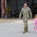 A U.S. Army soldier in OCP (Operational Camouflage Pattern) uniform walks hand-in-hand with a young girl dressed entirely in pink on a military airfield tarmac. The setting appears to be a helicopter maintenance hangar facility, with military rotary-wing aircraft visible in the background. The scene suggests a family reunion or visitation event at a military installation.