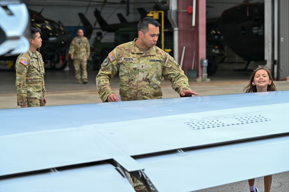A U.S. Army soldier named Gonzalez is explaining or demonstrating an aircraft component, likely a wing or rotor blade, to a young smiling girl at what appears to be a military aviation facility or hangar. Additional soldiers in OCP (Operational Camouflage Pattern) uniforms are visible in the background near helicopter hangars. The scene appears to be a family day, open house, or educational tour event.