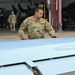A U.S. Army soldier named Gonzalez is explaining or demonstrating an aircraft component, likely a wing or rotor blade, to a young smiling girl at what appears to be a military aviation facility or hangar. Additional soldiers in OCP (Operational Camouflage Pattern) uniforms are visible in the background near helicopter hangars. The scene appears to be a family day, open house, or educational tour event.
