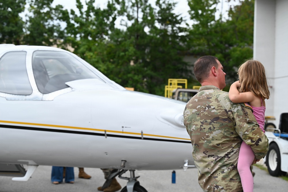 A military service member in OCP (Operational Camouflage Pattern) uniform holds a young child while standing near a small white civilian aircraft with yellow and black accent stripes. The scene appears to take place on an airport ramp or tarmac area, with trees and a hangar visible in the background. The moment suggests a family reunion or pre-departure interaction near the aircraft.
