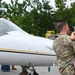 A military service member in OCP (Operational Camouflage Pattern) uniform holds a young child while standing near a small white civilian aircraft with yellow and black accent stripes. The scene appears to take place on an airport ramp or tarmac area, with trees and a hangar visible in the background. The moment suggests a family reunion or pre-departure interaction near the aircraft.
