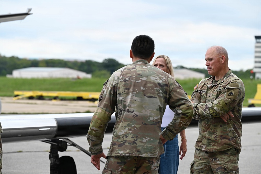 Three individuals are gathered on a military flight line or tarmac near an aircraft. Two are wearing U.S. Army OCP (Operational Camouflage Pattern) uniforms, while one civilian woman in casual attire stands between them. The senior uniformed individual on the right displays a name tape reading 'BLANCHARD' and appears to hold a general officer rank based on visible insignia.