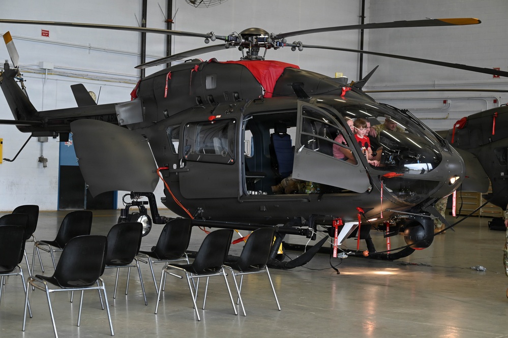 A dark gray military helicopter, likely a UH-72 Lakota or similar variant, is displayed inside a hangar with its doors open. Civilians, including what appears to be a child and adults, are seated inside the cockpit area, suggesting a public open house or family day event. Black folding chairs are arranged in front of the aircraft, further indicating an organized public viewing event.