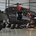 A dark gray military helicopter, likely a UH-72 Lakota or similar variant, is displayed inside a hangar with its doors open. Civilians, including what appears to be a child and adults, are seated inside the cockpit area, suggesting a public open house or family day event. Black folding chairs are arranged in front of the aircraft, further indicating an organized public viewing event.