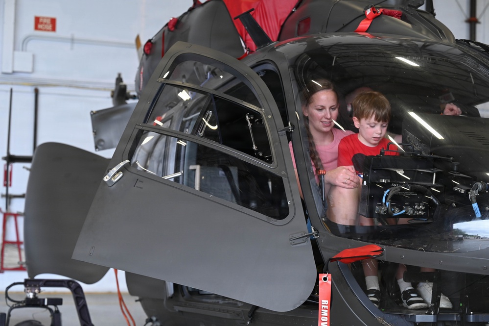 A woman and a young child are seated inside the cockpit of a military helicopter, likely during an open house or public display event at an aviation facility. The helicopter appears to be a dark gray/black military rotorcraft, possibly an AH-6 or similar light attack helicopter, parked inside a hangar. The child and adult appear to be exploring the cockpit instrumentation.